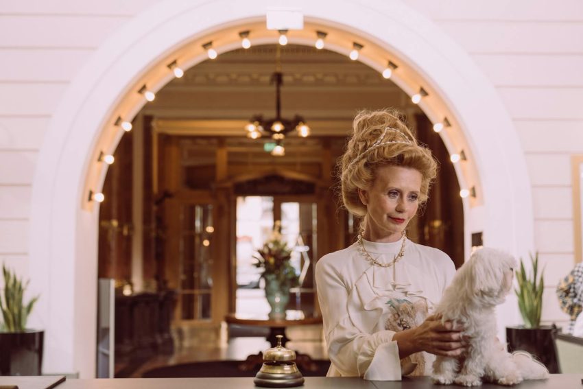 An elegant woman holding a dog at a luxurious hotel reception desk.